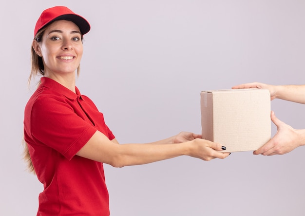 Sorridente giovane donna graziosa di consegna in uniforme dà cardbox a qualcuno che guarda davanti isolato sul muro bianco