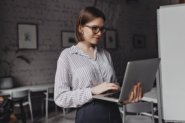 Sorridente giovane donna in camicetta a righe e occhiali cerchiati di nero guarda lo schermo del laptop su sfondo bianco bordo.