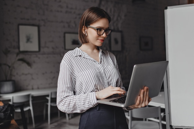 Sorridente giovane donna in camicetta a righe e occhiali cerchiati di nero guarda lo schermo del laptop su sfondo bianco bordo.