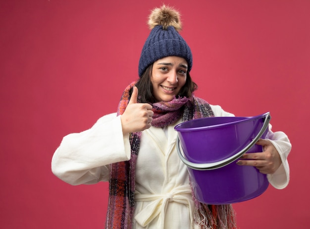 Sorridente giovane donna malata che indossa un abito invernale cappello e sciarpa tenendo il secchio di plastica che mostra il pollice in alto guardando la parte anteriore isolata sul muro rosa