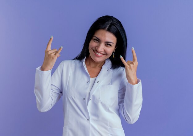 Sorridente giovane donna medico indossa abito medico cercando facendo segno di roccia