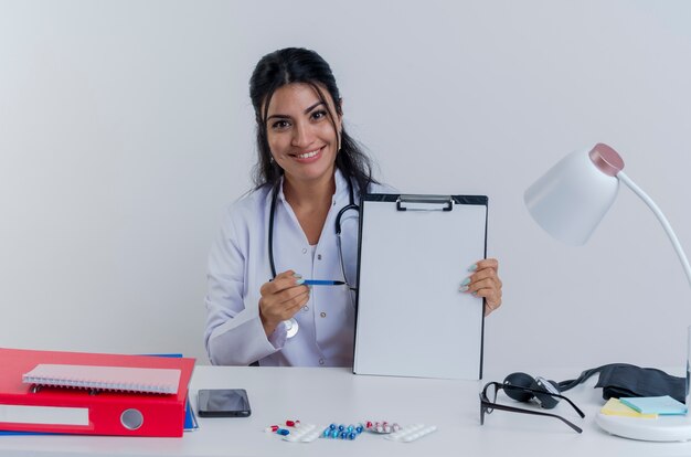 Sorridente giovane donna medico indossando abito medico e uno stetoscopio seduto alla scrivania con strumenti medici cercando che mostra appunti puntando con la penna a esso isolato