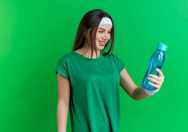 Sorridente giovane donna sportiva che indossa la fascia e braccialetti tenendo e guardando la bottiglia d'acqua