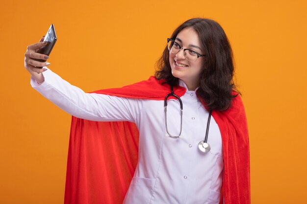 Sorridente giovane donna supereroe in mantello rosso che indossa l'uniforme del medico e lo stetoscopio con gli occhiali che prendono selfie isolato sulla parete arancione