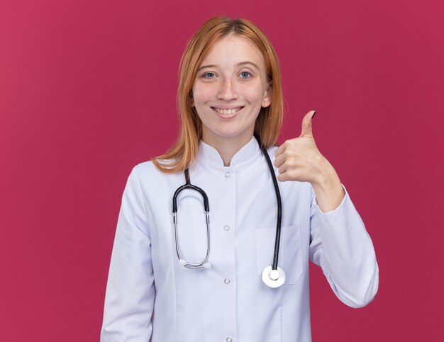 Sorridente giovane dottoressa allo zenzero che indossa una tunica medica e uno stetoscopio che mostra il pollice in su