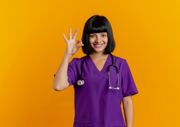 Sorridente giovane dottoressa bruna in uniforme con il segno giusto della mano di gesti dello stetoscopio