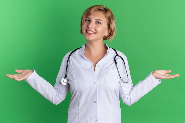 Sorridente giovane dottoressa che indossa una tunica medica con uno stetoscopio che allarga le mani isolate su sfondo verde