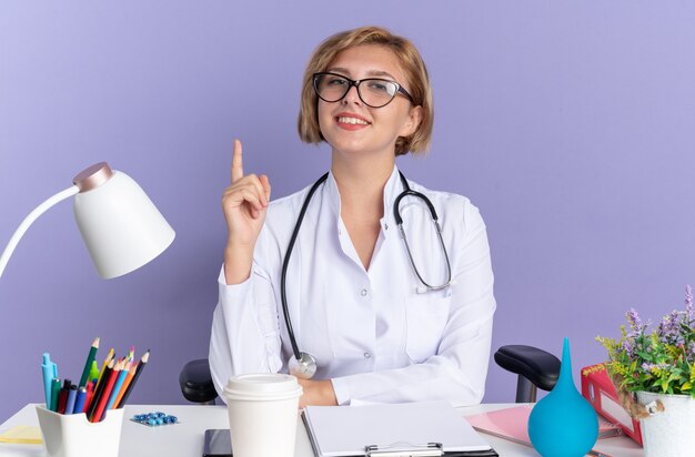 Sorridente giovane dottoressa che indossa una veste medica con stetoscopio e occhiali si siede al tavolo con strumenti medici puntati verso l'alto isolato su sfondo blu