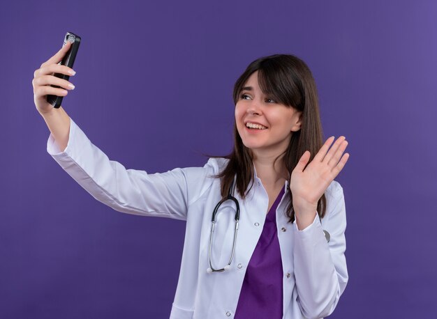 Sorridente giovane dottoressa in abito medico con lo stetoscopio guarda il telefono e gesti ciao su sfondo viola isolato