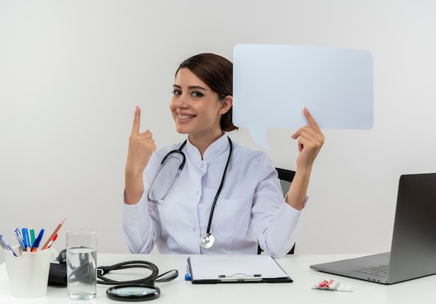 Sorridente giovane dottoressa indossa veste medica con stetoscopio seduto alla scrivania lavora sul computer con strumenti medici tenendo la bolla di chat poiunts fino a con lo spazio della copia