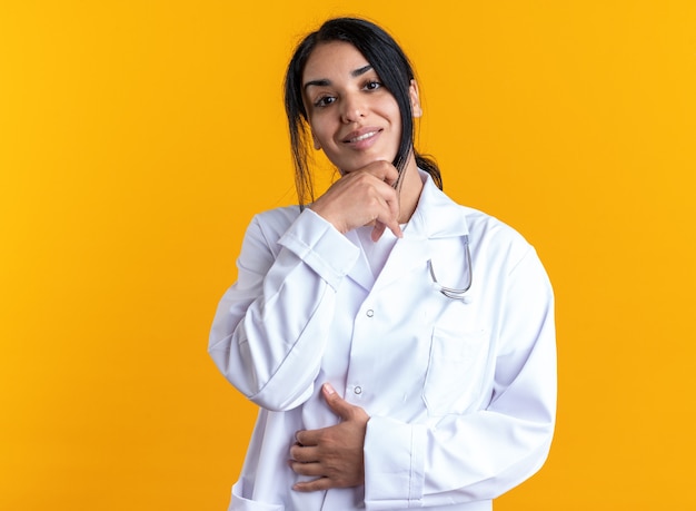 Sorridente giovane dottoressa indossando accappatoio medico con stetoscopio afferrato il mento isolato su sfondo giallo
