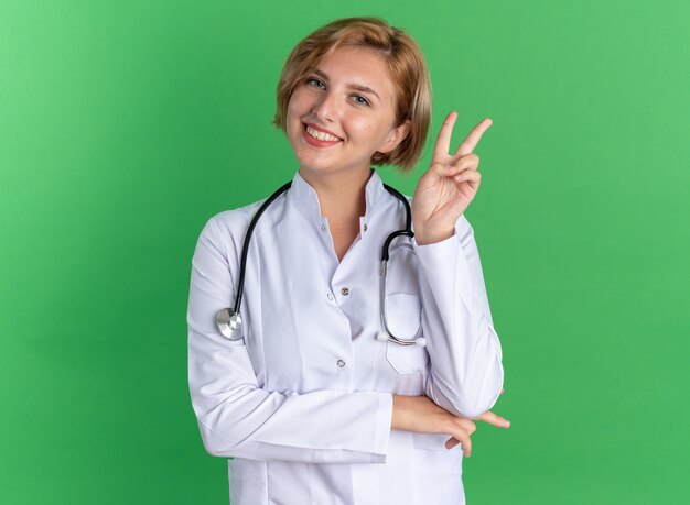 Sorridente giovane dottoressa indossando accappatoio medico con stetoscopio che mostra gesto di pace isolato su sfondo verde