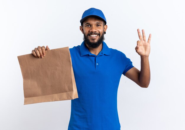 Sorridente giovane fattorino afro-americano che tiene in mano un pacchetto di cibo e gesticola segno ok isolato su sfondo bianco con spazio di copia copy