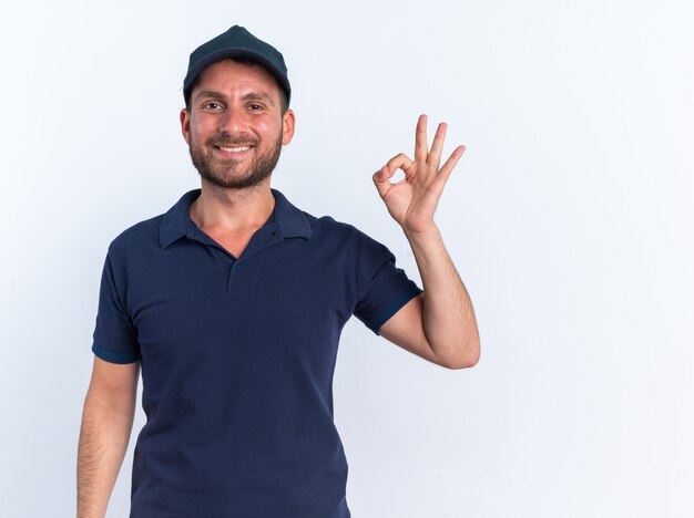 Sorridente giovane fattorino caucasico in uniforme blu e berretto che guarda l'obbiettivo facendo segno ok isolato sul muro bianco con copia spazio