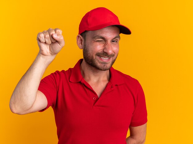 Sorridente giovane fattorino caucasico in uniforme rossa e berretto che guarda l'obbiettivo che fa l'occhiolino facendo un gesto di bussare isolato sul muro arancione