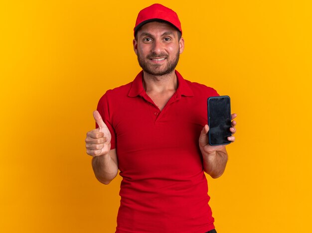 Sorridente giovane fattorino caucasico in uniforme rossa e berretto che guarda l'obbiettivo che mostra il telefono cellulare e il pollice in alto isolato sul muro arancione
