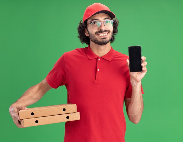 Sorridente giovane fattorino caucasico in uniforme rossa e berretto con gli occhiali che tengono i pacchetti di pizza che mostrano il telefono cellulare