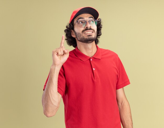 Sorridente giovane fattorino caucasico in uniforme rossa e berretto con gli occhiali guardando il lato rivolto verso l'alto isolato sulla parete verde oliva con spazio di copia