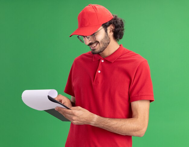 Sorridente giovane fattorino caucasico in uniforme rossa e berretto con gli occhiali in piedi nella vista di profilo tenendo e guardando gli appunti che puntano il dito su di esso