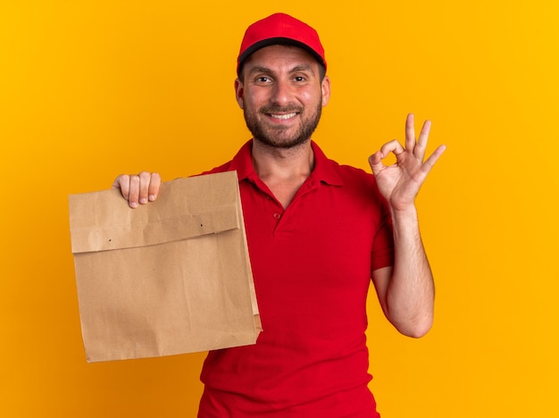 Sorridente giovane fattorino caucasico in uniforme rossa e berretto con in mano un pacchetto di carta guardando la telecamera facendo segno ok isolato sul muro arancione