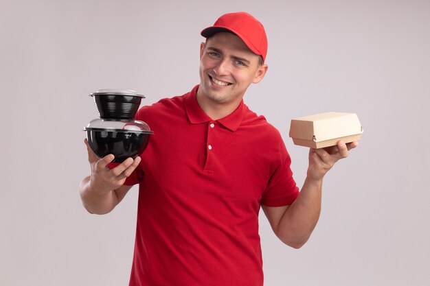 Sorridente giovane fattorino che indossa l'uniforme con cappuccio che tiene contenitori per alimenti con pacchetto di carta alimentare isolato sulla parete bianca