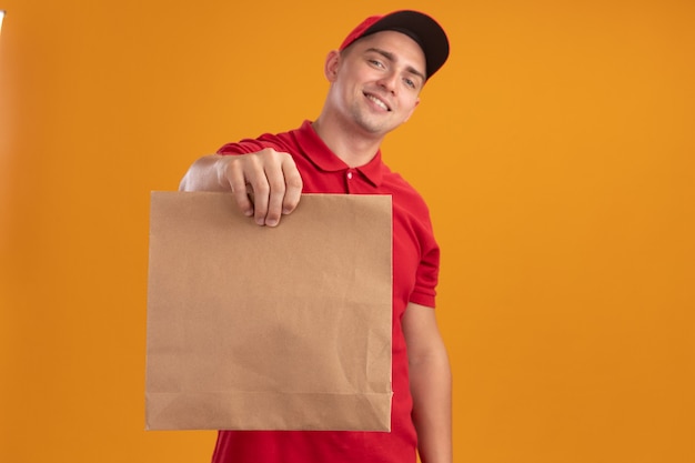 Sorridente giovane fattorino che indossa l'uniforme con cappuccio che tiene fuori il pacchetto di carta alimentare nella parte anteriore isolata sulla parete arancione