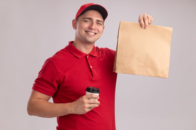 Sorridente giovane fattorino che indossa l'uniforme con cappuccio che tiene il pacchetto di cibo di carta con una tazza di caffè isolato sulla parete bianca