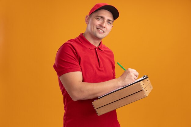 Sorridente giovane fattorino che indossa l'uniforme con il cappuccio che tiene le scatole della pizza e scrive qualcosa negli appunti isolati sulla parete arancione