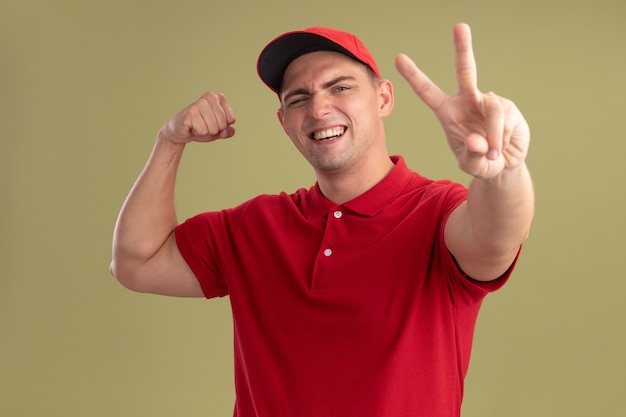 Sorridente giovane fattorino che indossa l'uniforme e cappuccio che mostra gesto di pace facendo un gesto forte isolato sulla parete verde oliva