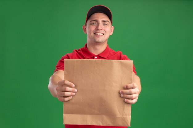 Sorridente giovane fattorino che indossa l'uniforme e cappuccio che tiene fuori il pacchetto di cibo di carta nella parte anteriore isolata sulla parete verde