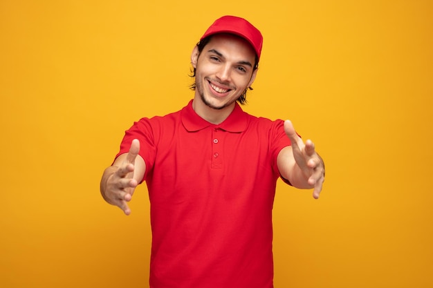 sorridente giovane fattorino che indossa uniforme e berretto guardando la fotocamera allungando le mani verso la fotocamera isolata su sfondo giallo