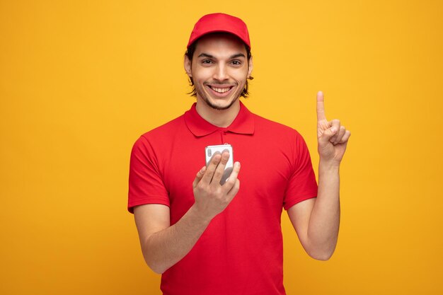 sorridente giovane fattorino che indossa uniforme e cappuccio che tiene il telefono cellulare guardando la fotocamera rivolta verso l'alto isolata su sfondo giallo