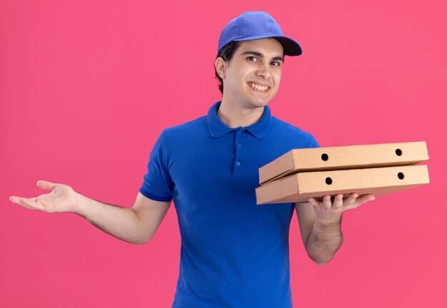 Sorridente giovane fattorino in uniforme blu e berretto che tiene i pacchetti di pizza guardando la parte anteriore che mostra la mano vuota isolata sul muro rosa