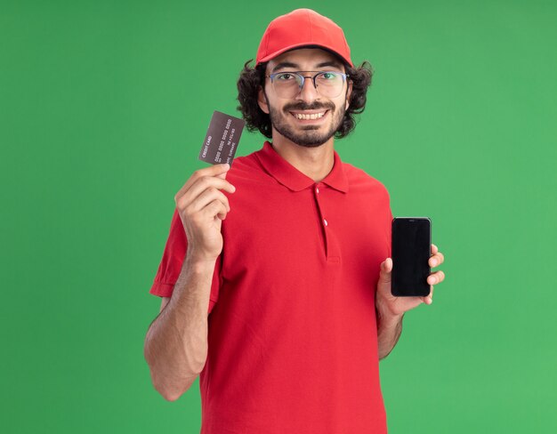 Sorridente giovane fattorino in uniforme rossa e berretto con gli occhiali che mostra il telefono cellulare e la carta di credito davanti guardando la parte anteriore isolata sulla parete verde con spazio di copia