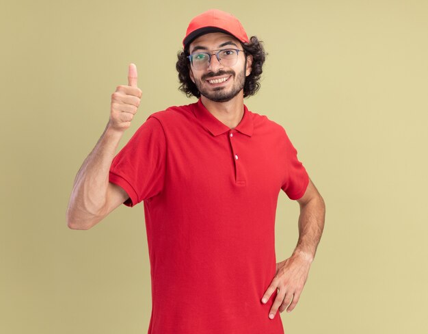 Sorridente giovane fattorino in uniforme rossa e berretto con gli occhiali guardando davanti tenendo la mano sulla vita che mostra pollice in su isolato sulla parete verde oliva con spazio di copia