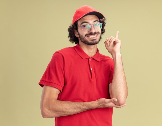 Sorridente giovane fattorino in uniforme rossa e berretto con gli occhiali guardando la parte anteriore rivolta verso l'alto isolata sulla parete verde oliva