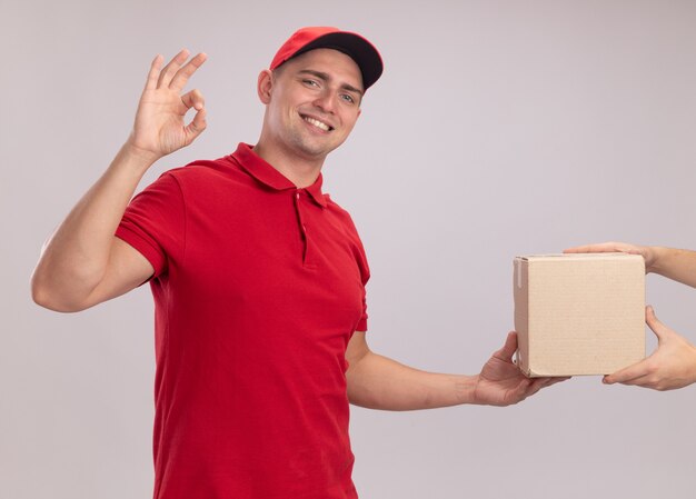 Sorridente giovane fattorino indossando l'uniforme con cappuccio che dà scatola al cliente che mostra gesto giusto isolato sulla parete bianca