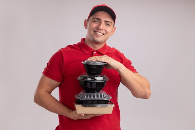 Sorridente giovane fattorino indossando l'uniforme con cappuccio che tiene contenitori per alimenti isolati sulla parete bianca