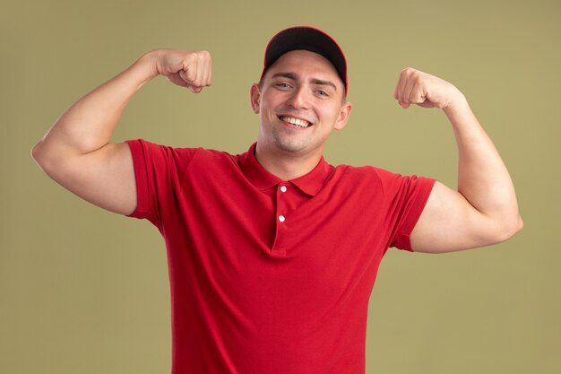 Sorridente giovane fattorino indossando l'uniforme e berretto che mostra un forte gesto isolato sulla parete verde oliva