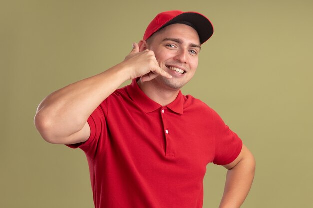 Sorridente giovane fattorino indossando l'uniforme e cappuccio che mostra il gesto di chiamata telefonica isolato sulla parete verde oliva