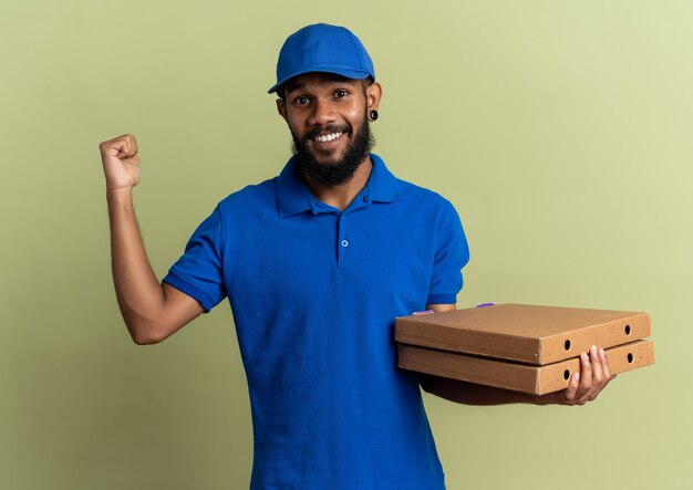 sorridente giovane fattorino tenendo scatole per pizza e puntando indietro isolato su parete verde oliva con spazio di copia