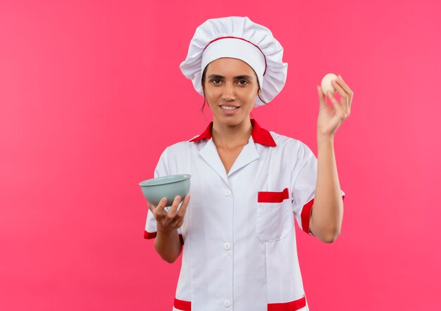 Sorridente giovane femmina cuoco indossando chef uniforme tenendo l'uovo e la ciotola con lo spazio della copia