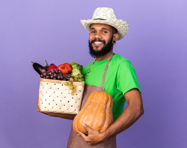 Sorridente giovane giardiniere afro-americano che indossa un cappello da giardinaggio che tiene un cesto di verdure con zucca isolata sulla parete blu