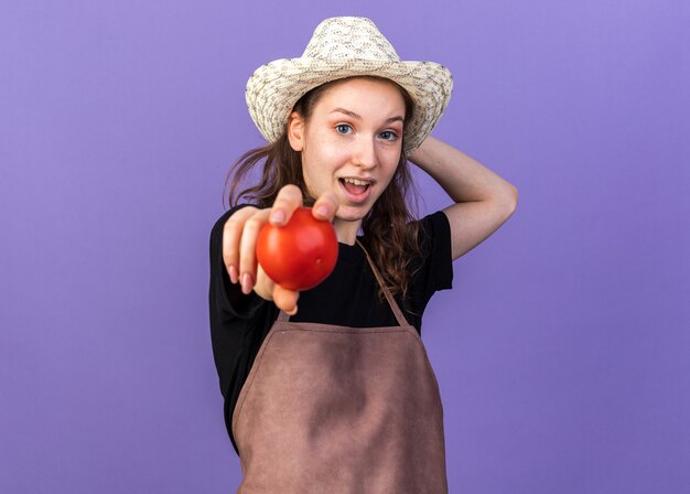 Sorridente giovane giardiniere femminile che indossa cappello da giardinaggio tenendo fuori il pomodoro isolato sulla parete blu