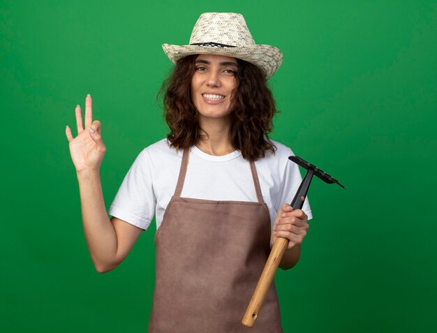 Sorridente giovane giardiniere femminile in uniforme che indossa cappello da giardinaggio che tiene il rastrello che mostra gesto giusto isolato sulla parete verde