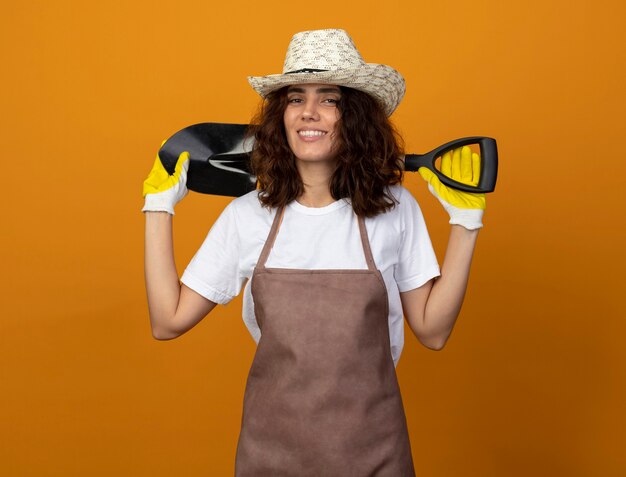 Sorridente giovane giardiniere femminile in uniforme che indossa cappello da giardinaggio e guanti che mettono la vanga dietro al collo