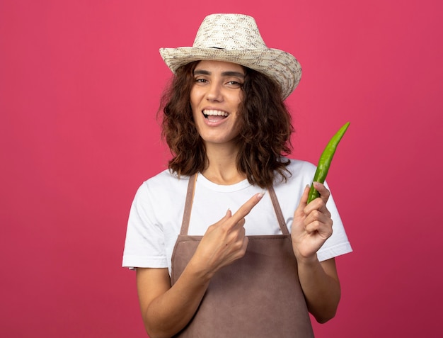 Sorridente giovane giardiniere femminile in uniforme che indossa cappello da giardinaggio holding e punti al pepe isolato sul colore rosa