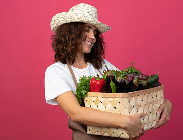 Sorridente giovane giardiniere femminile in uniforme che indossa cappello da giardinaggio tenendo e guardando il cesto di verdure