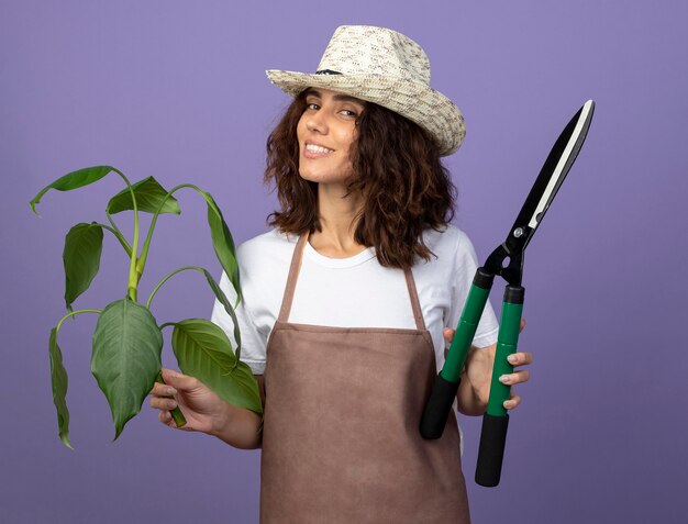 Sorridente giovane giardiniere femminile in uniforme che indossa cappello da giardinaggio tenendo la pianta della holding della pianta con i clippers