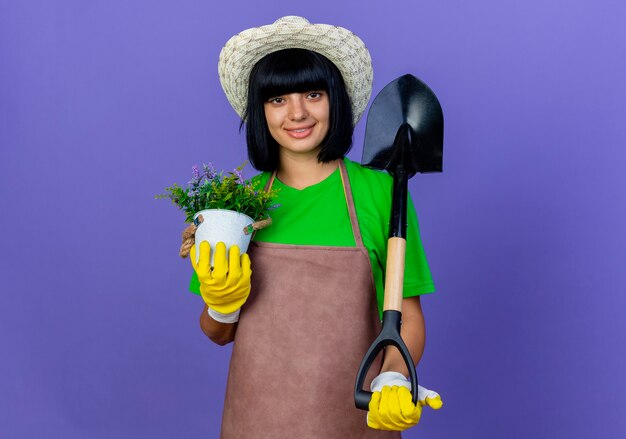 Sorridente giovane giardiniere femminile in uniforme che indossa cappello e guanti da giardinaggio tiene la vanga